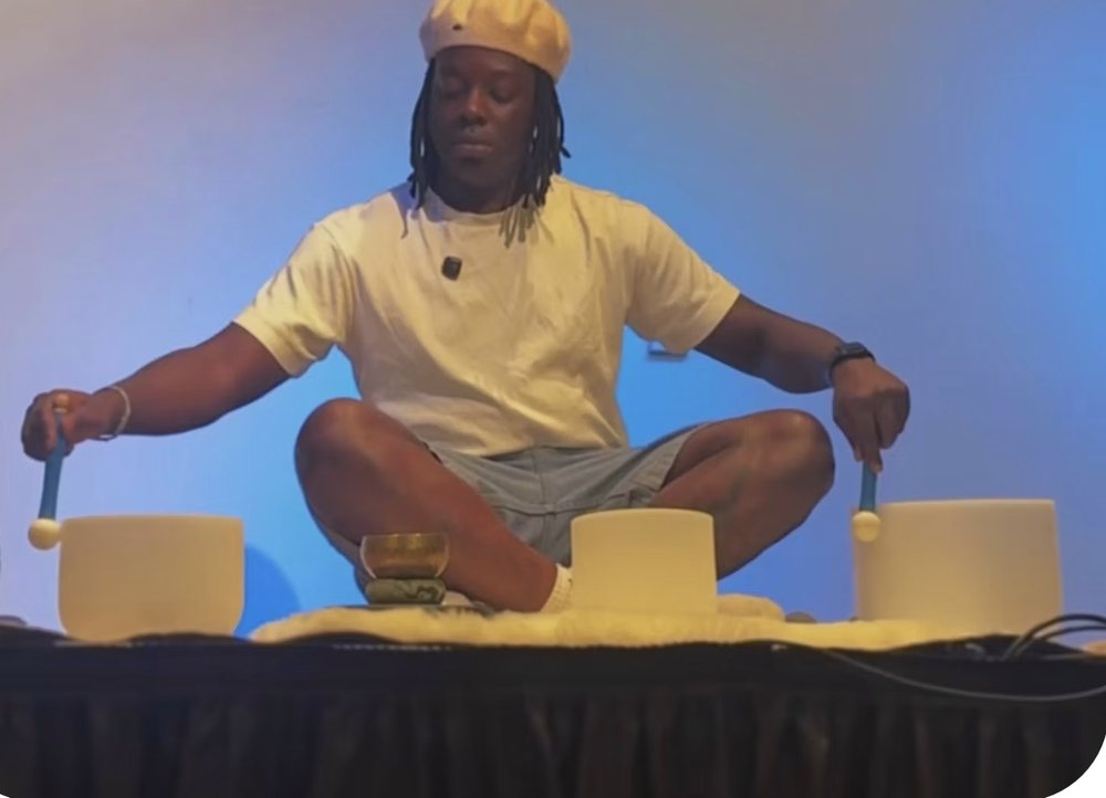 Raymell sitting cross-legged with three singing bowls on a stage lit in blue