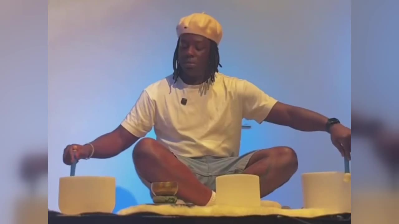 Raymell sitting cross-legged with three singing bowls on a stage lit in blue